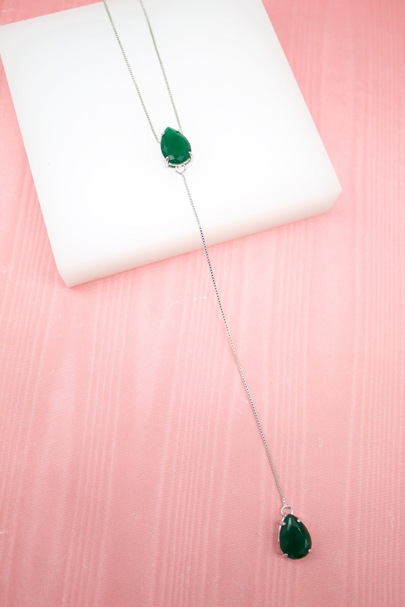 A delicate silver y-necklace features two teardrop green stones against a white and pink background.