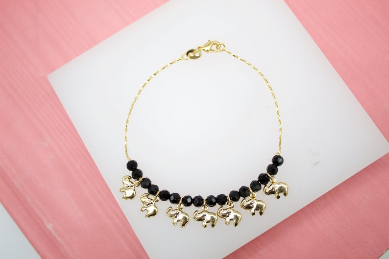 A delicate gold chain bracelet features black beaded accents and dangling gold elephant charms, displayed on a white stand against a pink background.