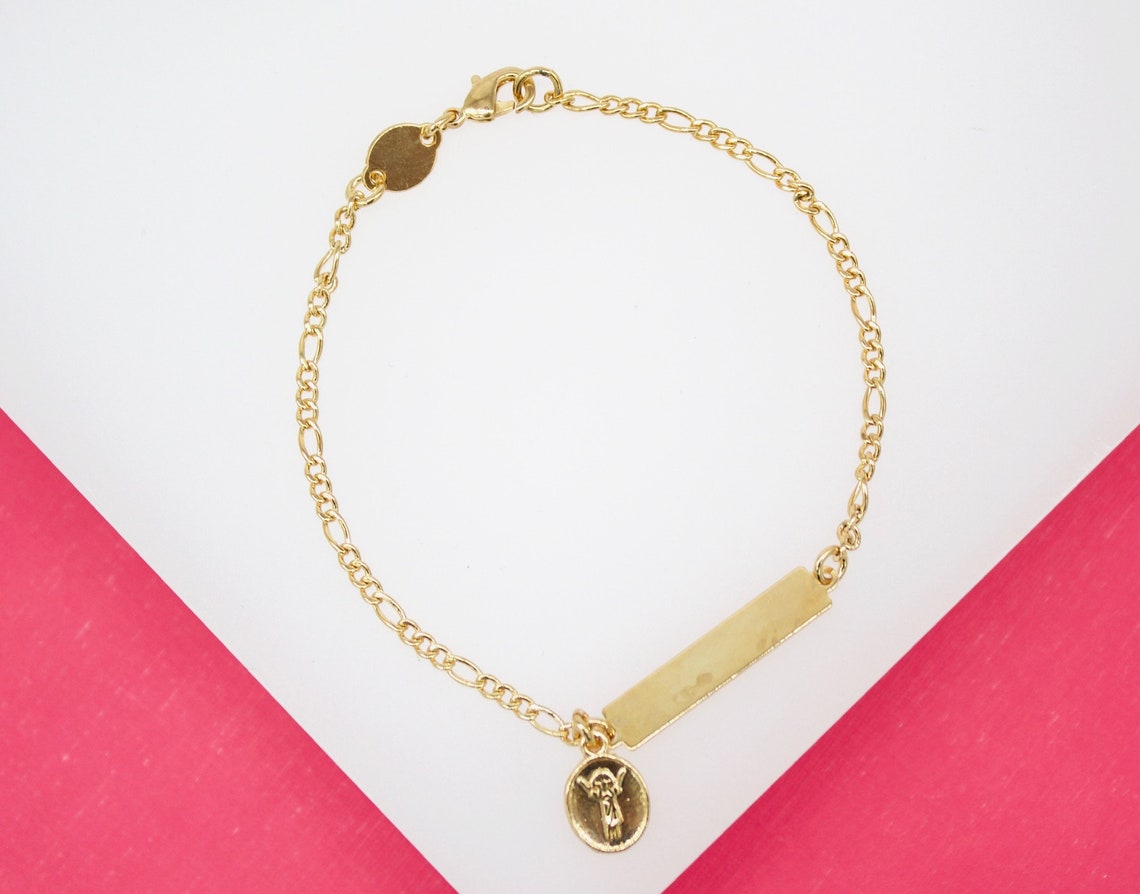 A delicate gold link bracelet rests on a white and pink background, featuring an ID tag and a small round angel medallion.