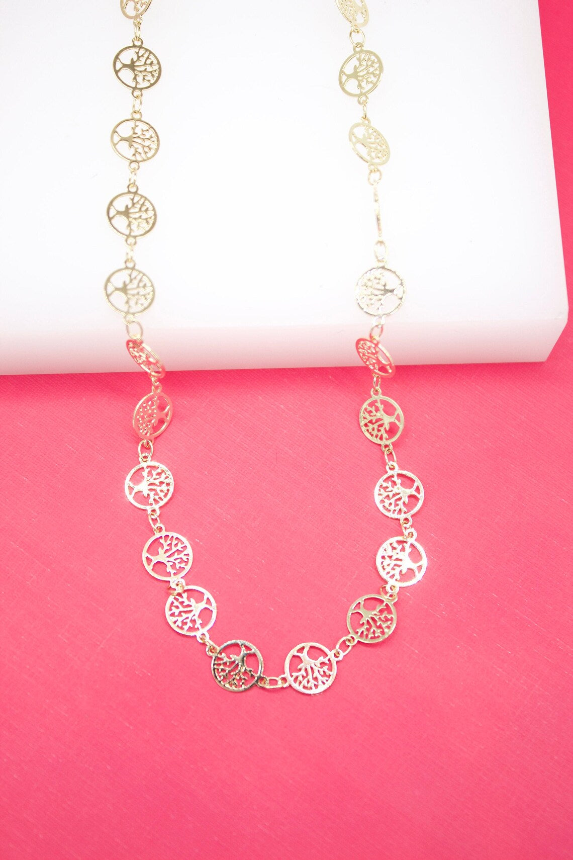 A gold Tree of Life charm necklace rests against a vibrant pink backdrop.