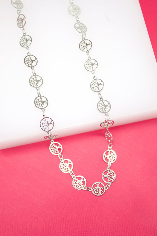 A delicate silver necklace featuring interconnected tree-of-life pendants rests gracefully on a white display surface against a vibrant pink background.