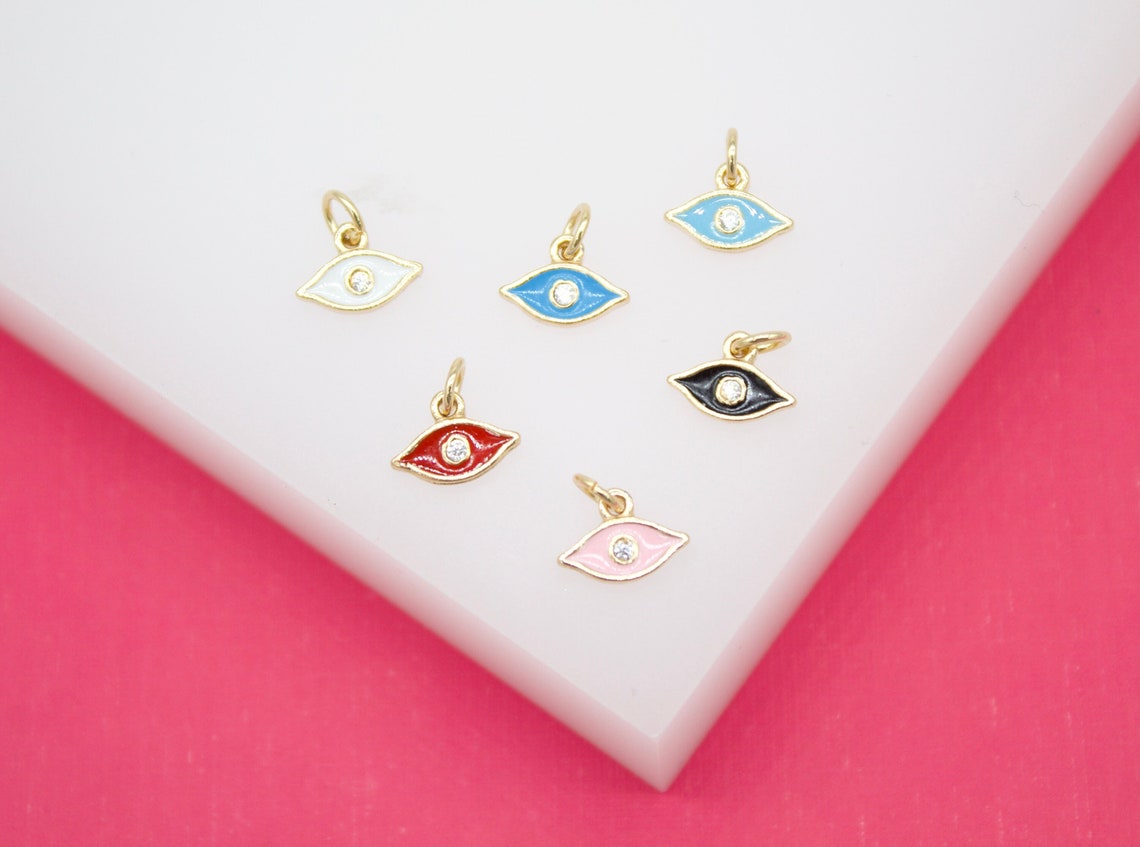 Six colorful enamel evil eye charms with gold settings rest on a white surface against a vibrant pink background.