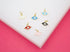 Six colorful enamel evil eye charms with gold settings rest on a white surface against a vibrant pink background.