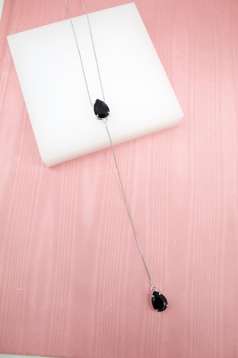 A delicate silver lariat necklace features two black teardrop-shaped gemstones resting on a white display block against a textured pink surface.