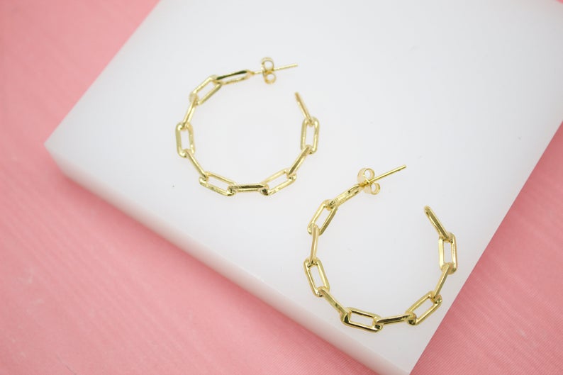 A pair of gold paperclip chain hoop earrings rests on a white display block against a pink background.