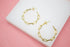 A pair of gold paperclip chain hoop earrings rests on a white display block against a pink background.