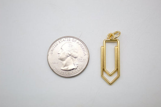 A gold paperclip charm rests beside a US quarter for size comparison on a plain surface.