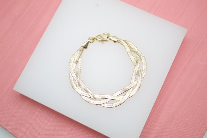 A gleaming gold-plated triple herringbone chain bracelet rests on a white display block against a textured pink background.