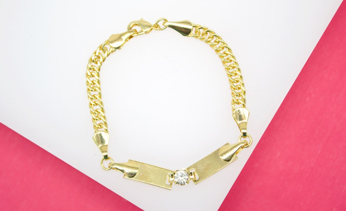 A gold curb chain bracelet featuring an ID plate centered with a sparkling diamond lies against a white and pink background.