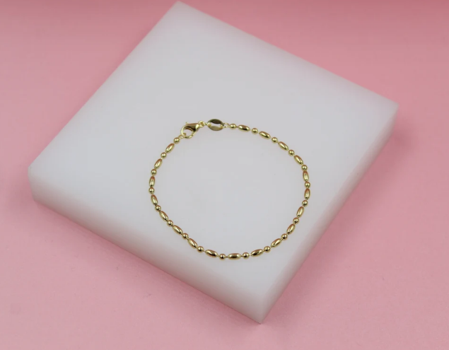 A delicate gold beaded bracelet rests on a white display block against a pink background.