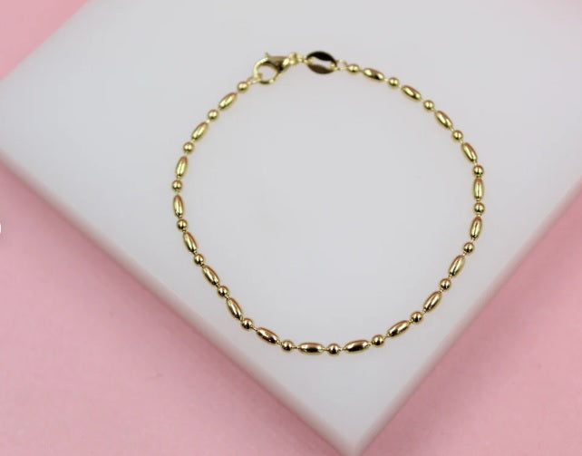 A delicate gold beaded bracelet rests on a white display surface against a pink background.