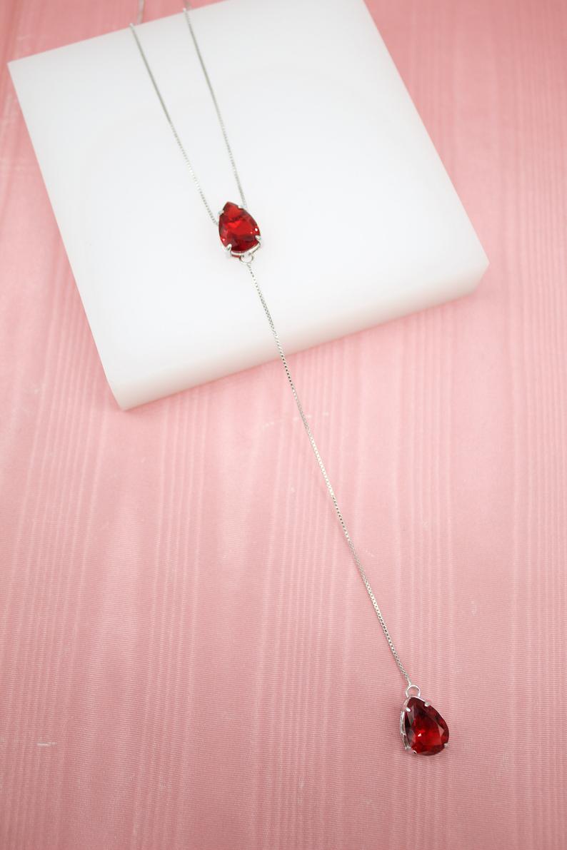 A silver lariat necklace featuring two teardrop-cut red gemstones rests on a white display against a pink background.