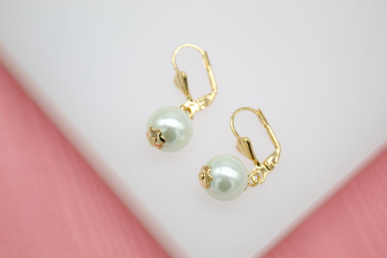 A pair of elegant gold leverback earrings feature dangling white pearl drops resting on a clean white and pink surface.