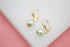 A pair of elegant gold leverback earrings feature dangling white pearl drops resting on a clean white and pink surface.