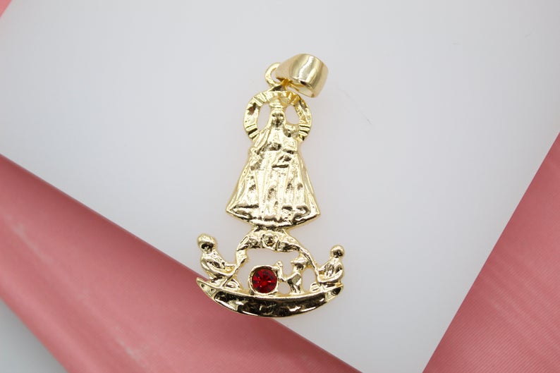 A shiny gold pendant features an intricate religious figure holding a baby above a crescent base accented with a sparkling red gemstone.