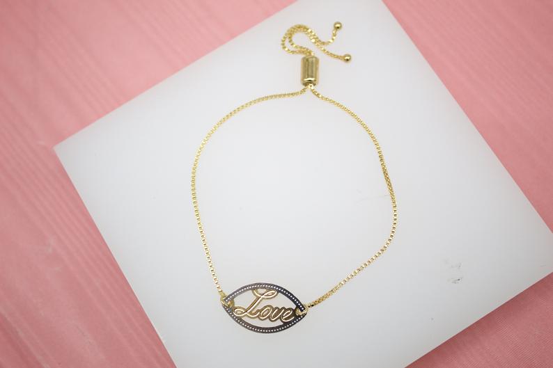 A gold slider bracelet featuring an evil eye charm with the word "Love" inscribed in cursive rests on a white display surface.