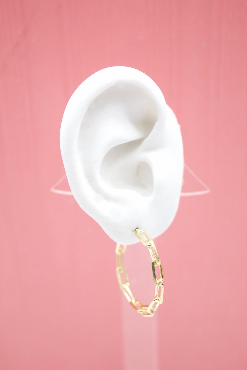 A white mannequin ear displays a gleaming gold chain-link hoop earring against a soft pink background.