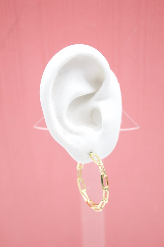 A white mannequin ear displays a gleaming gold chain-link hoop earring against a soft pink background.