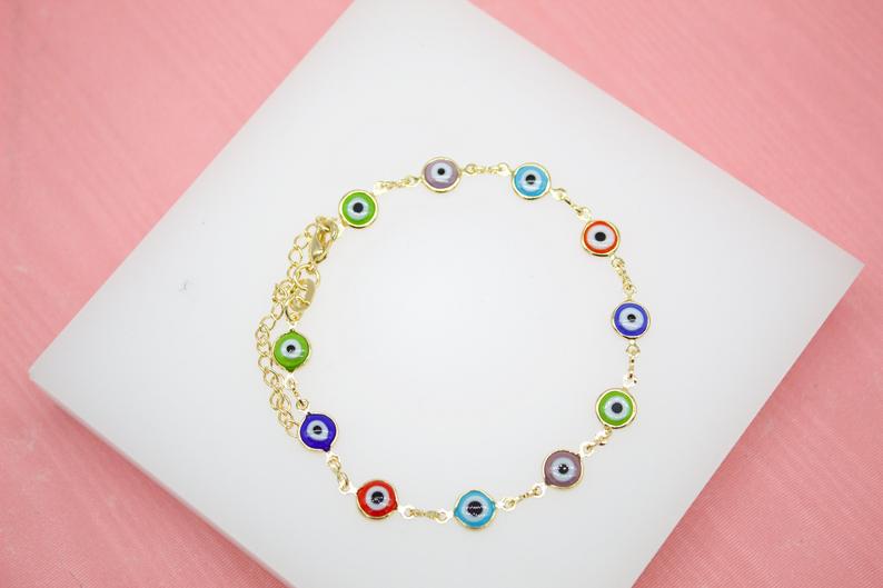 A gold chain bracelet featuring colorful evil eye charms rests on a white display against a pink background.