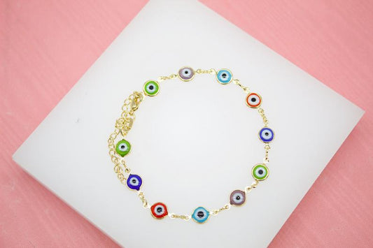 A gold chain bracelet featuring colorful evil eye charms rests on a white display against a pink background.