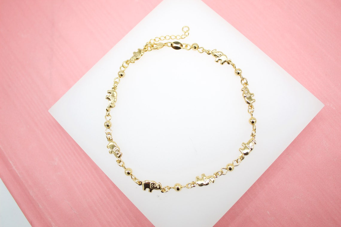 A delicate gold chain bracelet features alternating tiny elephant charms and small beads, resting on a white display against a pink background.