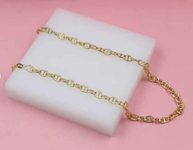 A glossy gold anchor chain necklace rests neatly on a white display block against a soft pink background.