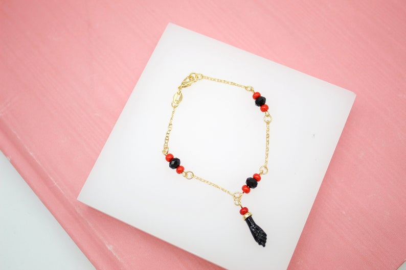 A delicate gold chain bracelet features black and red beads and a figa charm, resting on a white display block against a pink background.