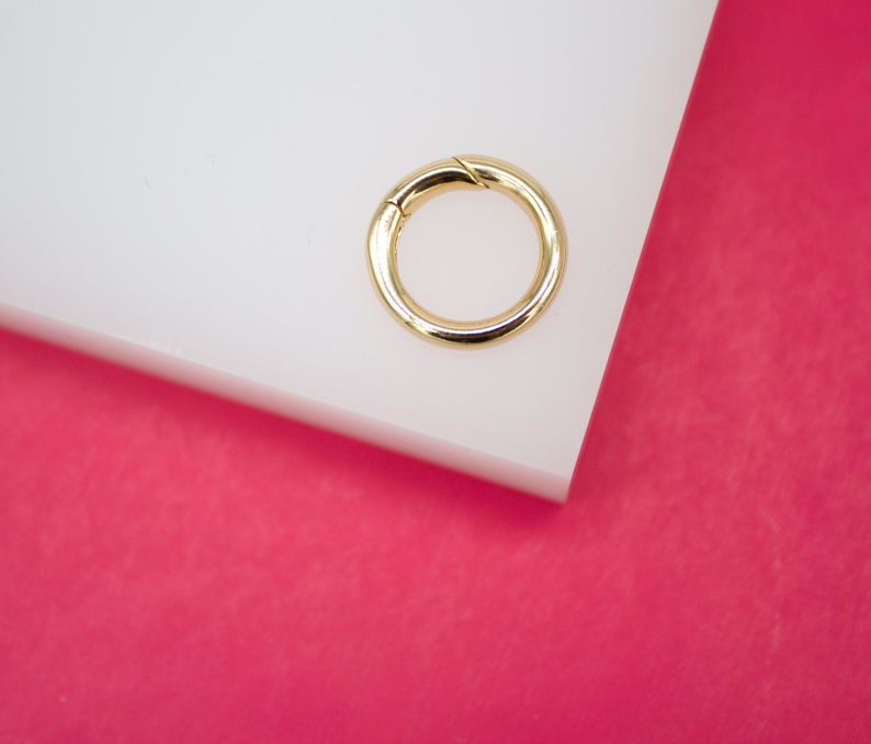 A polished gold clicker ring rests on a white display against a vibrant pink background.