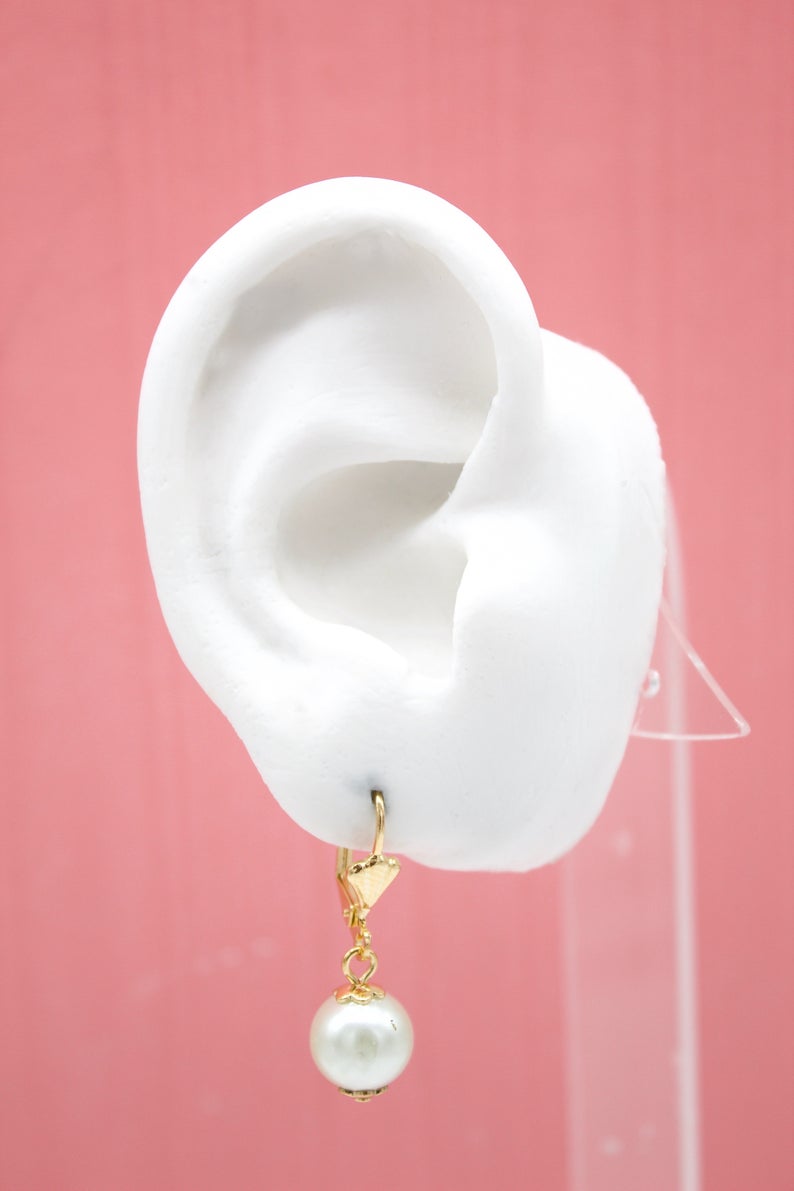 A white mannequin ear displays a gold leverback earring featuring a dangling white pearl against a solid pink background.