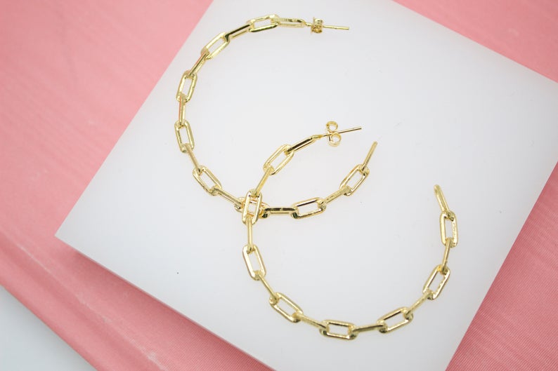 A pair of shiny gold paperclip chain hoop earrings rests on a white display surface against a pink background.