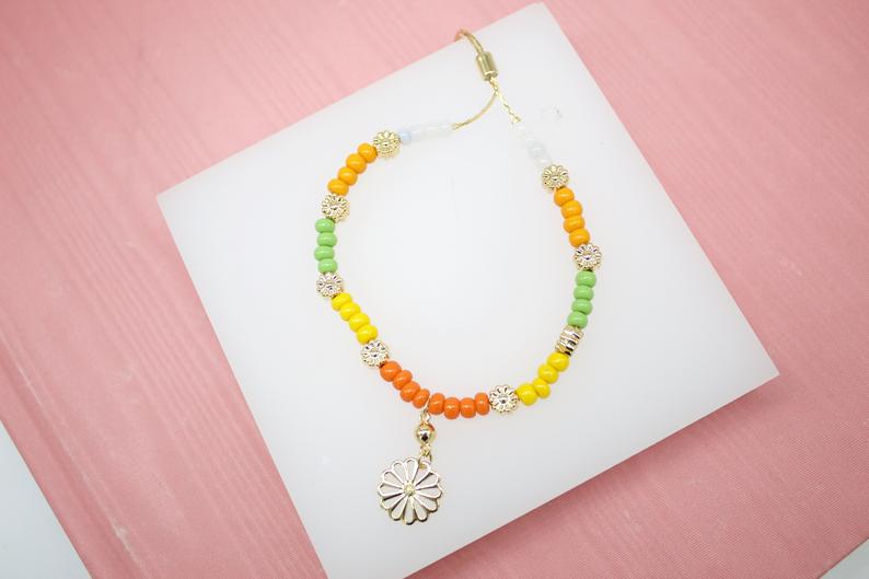 A colorful beaded slider bracelet featuring green, yellow, and orange seed beads and a delicate daisy charm rests on a white display block.