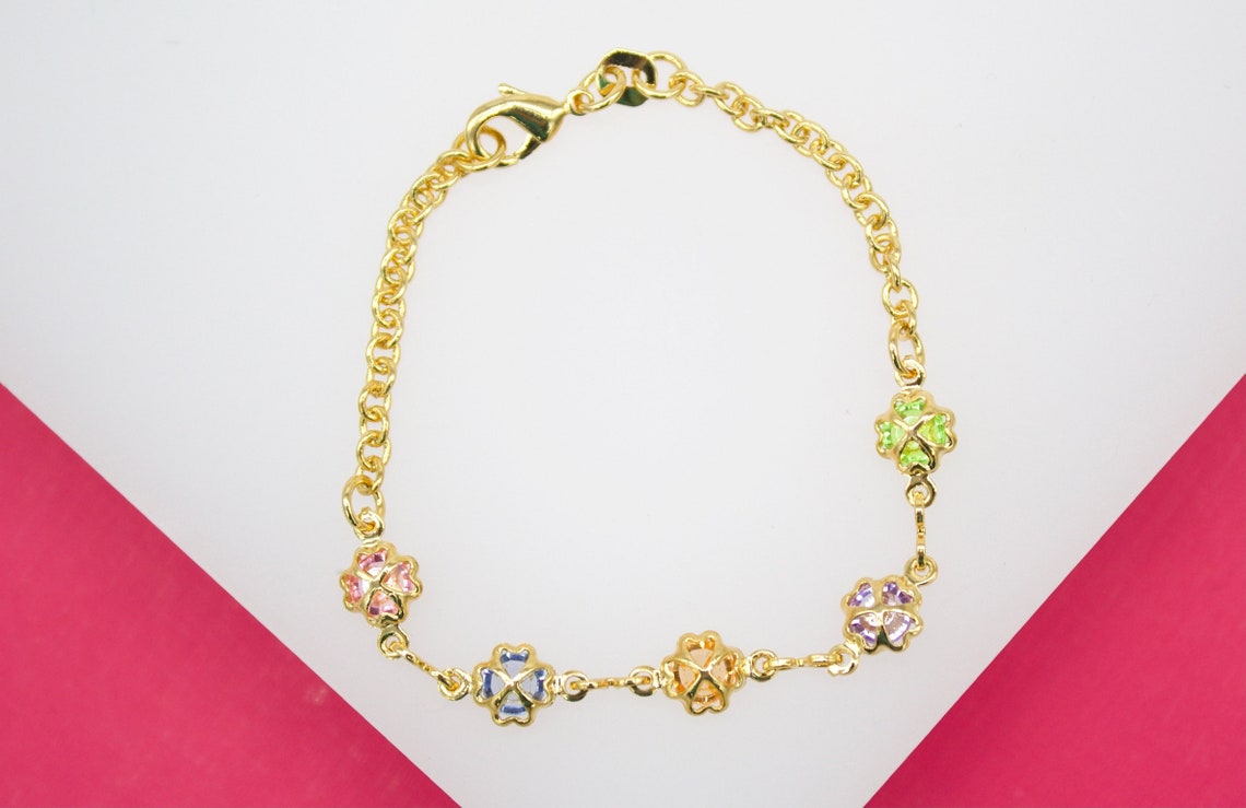 A delicate gold chain bracelet features colorful crystal four-leaf clover charms resting on a white and pink background.