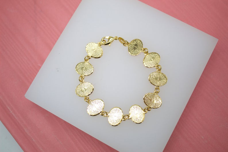 A delicate gold-plated link bracelet featuring multiple textured oval medals lies displayed on a white platform against a pink background.
