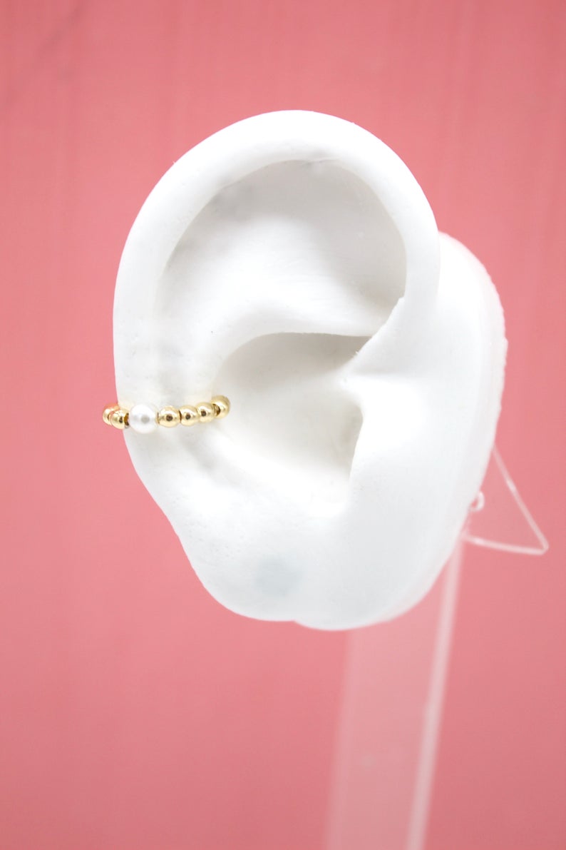 A white display ear model showcases a delicate gold beaded ear cuff featuring a central white pearl.
