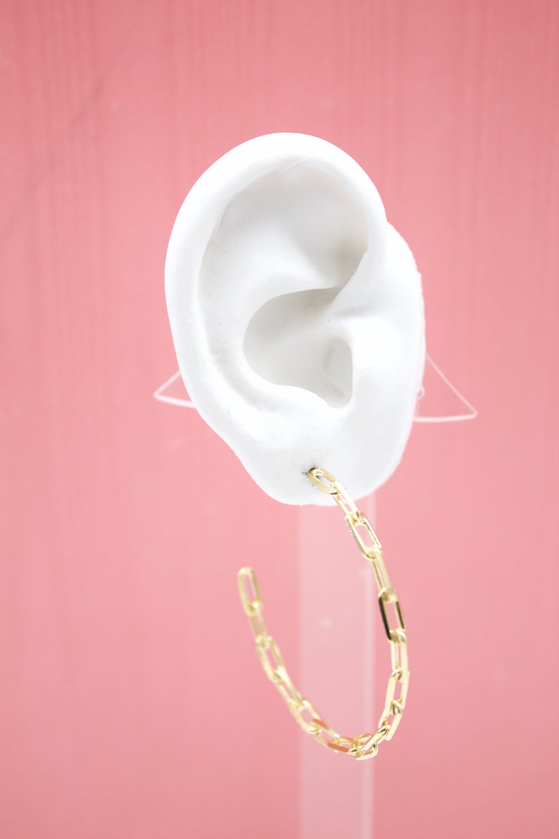 A white mannequin ear displays a stylish gold paperclip chain hoop earring against a vibrant pink background.