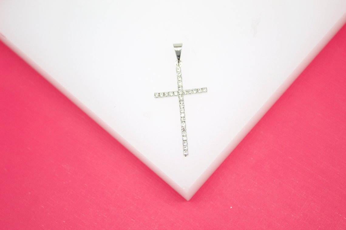 A silver cross pendant encrusted with sparkling rhinestones rests on a white display against a vibrant pink background.