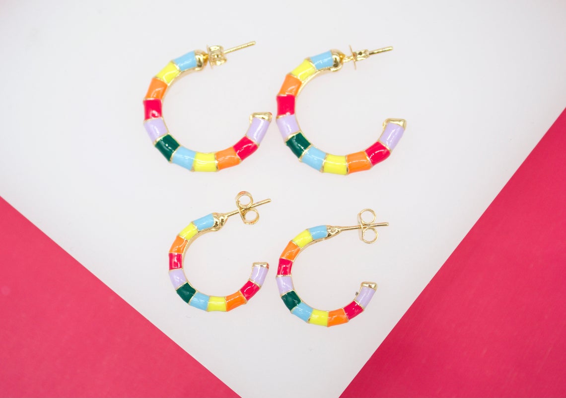 Four vibrant gold hoop earrings with multi-colored enamel segments rest against a pink and white background.