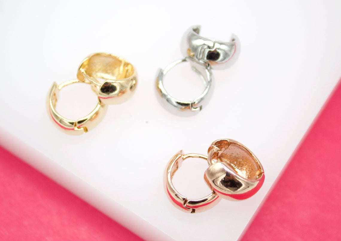 Three pairs of small, chunky huggie hoop earrings in gold, silver, and rose gold rest neatly on a white display surface against a vibrant pink background.