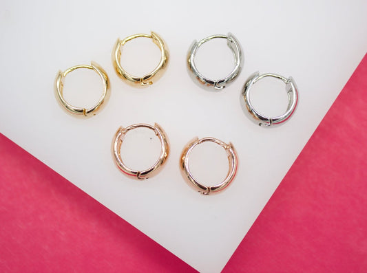 Three pairs of small hoop earrings in gold, silver, and rose gold finishes rest neatly on a white display surface against a vibrant pink background.