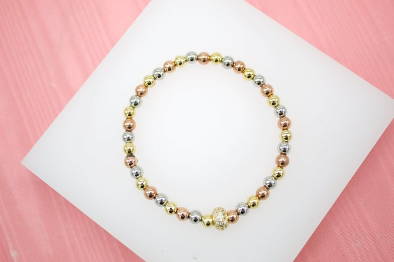 A tri-color beaded stretch bracelet featuring alternating yellow gold, rose gold, and silver spheres rests on a white display surface.