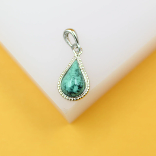 A teardrop-shaped turquoise gemstone pendant with a silver beaded setting rests on a white surface against an orange background.
