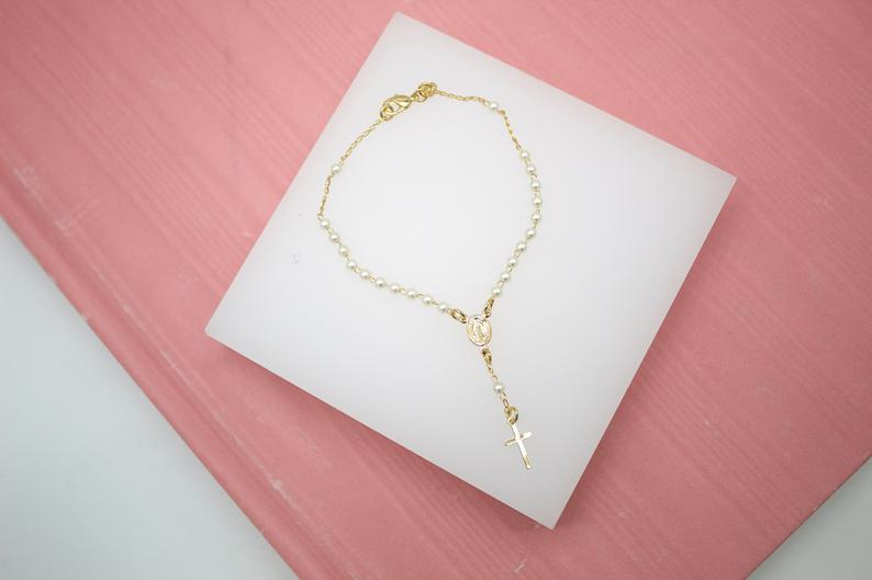 A delicate gold rosary bracelet featuring small white pearls and a tiny crucifix lies displayed on a white platform against a pink wooden background.