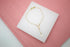 A delicate gold rosary bracelet featuring small white pearls and a tiny crucifix lies displayed on a white platform against a pink wooden background.