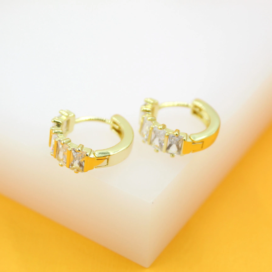 Two gold hoop earrings featuring sparkling baguette-cut cubic zirconia stones rest on a white surface against an orange background.