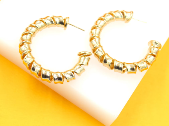 A pair of glossy gold spiral hoop earrings rests on a white block against a bright yellow background.