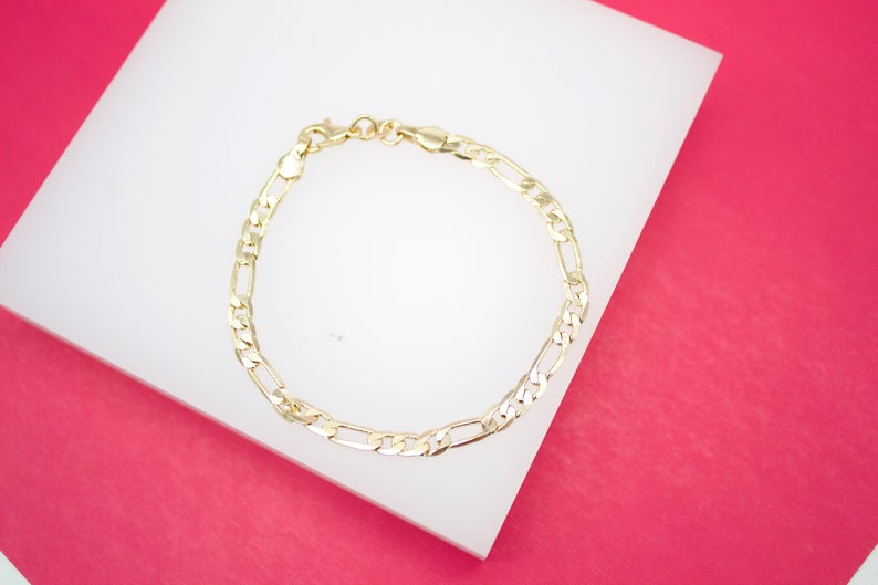A delicate gold Figaro link chain bracelet rests on a white display against a vibrant pink background.
