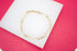 A delicate gold Figaro link chain bracelet rests on a white display against a vibrant pink background.