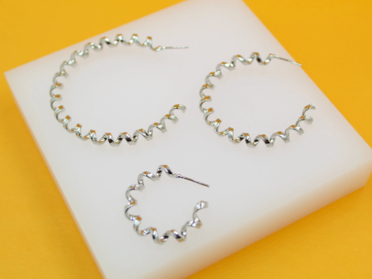 Three silver spiral hoop earrings of varying sizes rest on a white display block against a vibrant yellow background.
