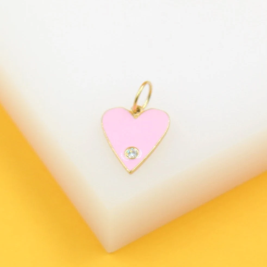 A gold heart-shaped pendant featuring pink enamel and a sparkling crystal accents rests on a white and yellow surface.