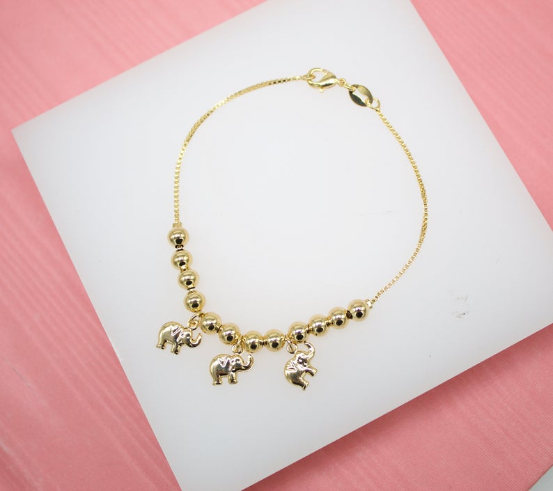 A delicate gold-plated chain bracelet features a row of shiny spherical beads and three dangling elephant charms on a white display surface.